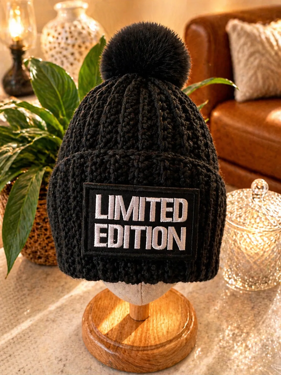 Black crocheted beanie with a black pom pom and white limited edition patch, displayed on a wooden stand in a warm, styled home setting