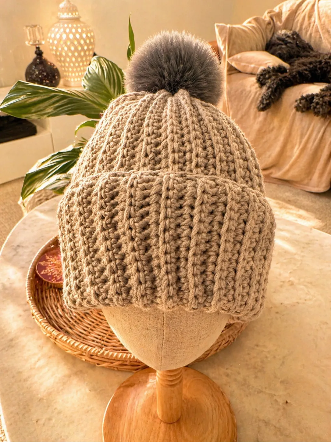 Oatmeal-coloured crocheted beanie with cable knit texture and charcoal grey pom pom, displayed on a wooden form with potted plants and home décor in the background