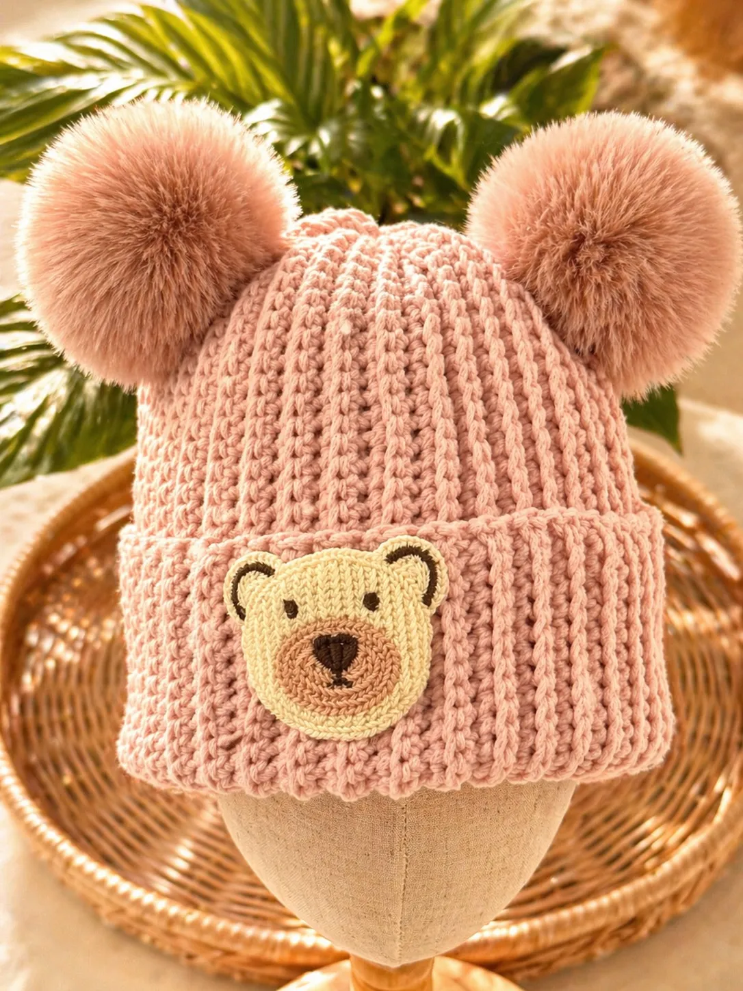 Blush pink crocheted baby beanie with two large fuzzy pom pom ears and a yellow embroidered bear face on the front, displayed on a stand with green plant behind it