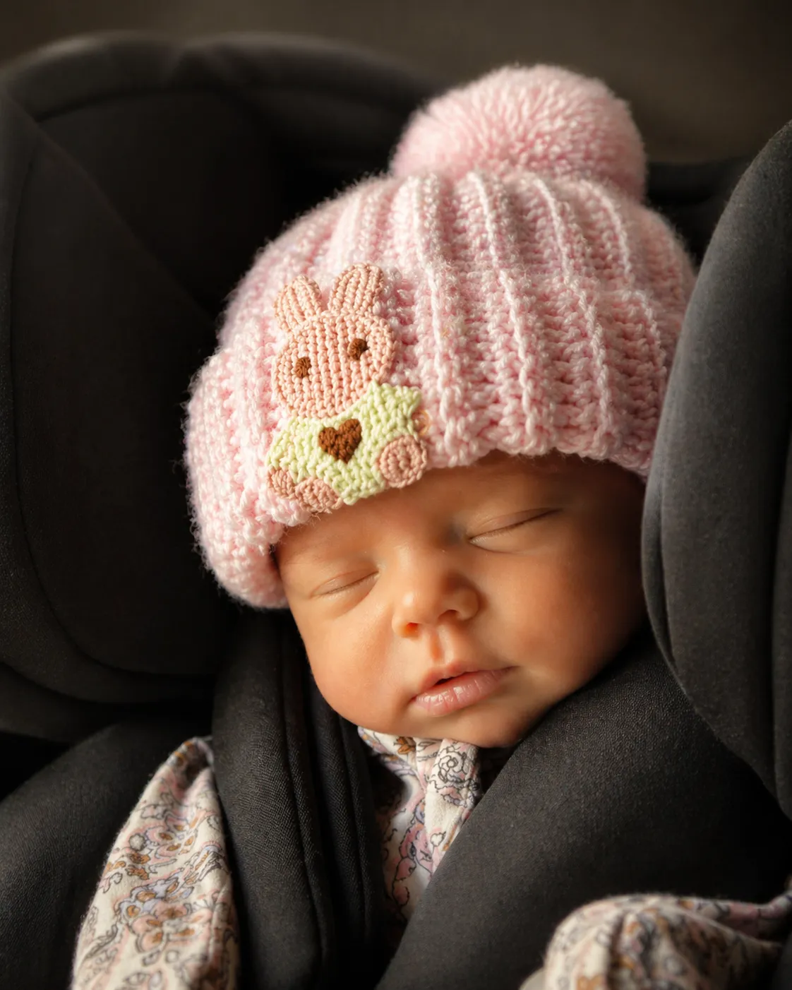 A newborn asleep in a car seat, wearing a soft pink ribbed beanie with a bunny patch and pink pom pom.