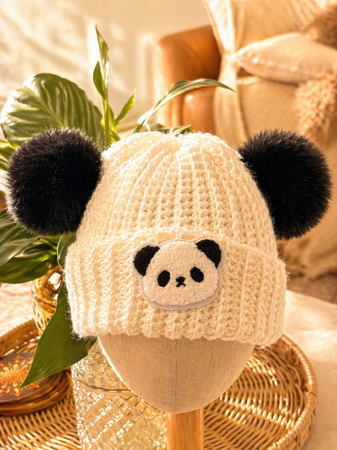 Hand-crocheted cream beanie with an embroidered panda face and two black fuzzy pom poms positioned as ears, displayed on a wooden stand with plant stems and woven basket details in the background