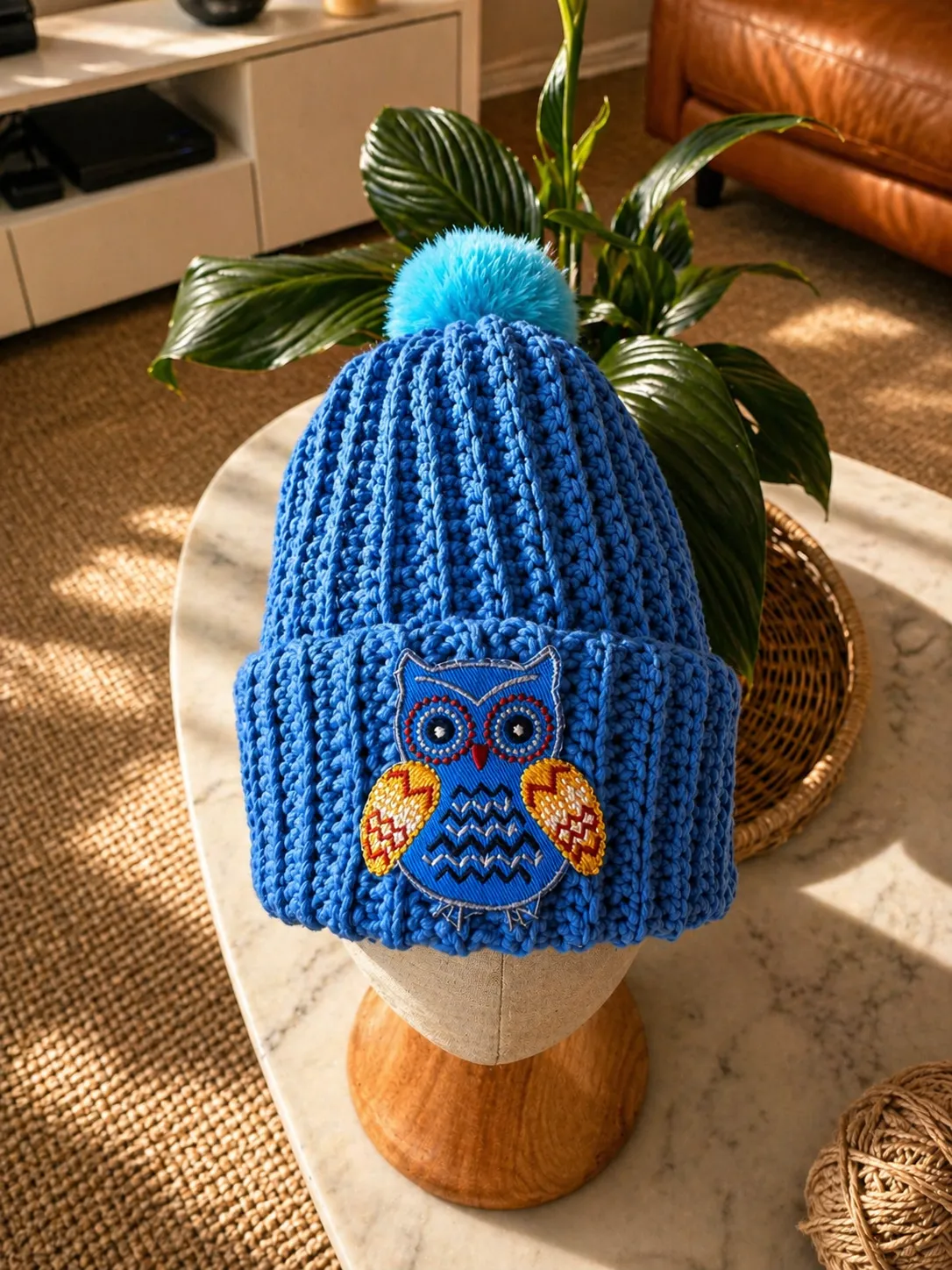 Hand-crocheted blue beanie with bright blue pom pom, featuring an embroidered owl patch with red eyes and gold wings