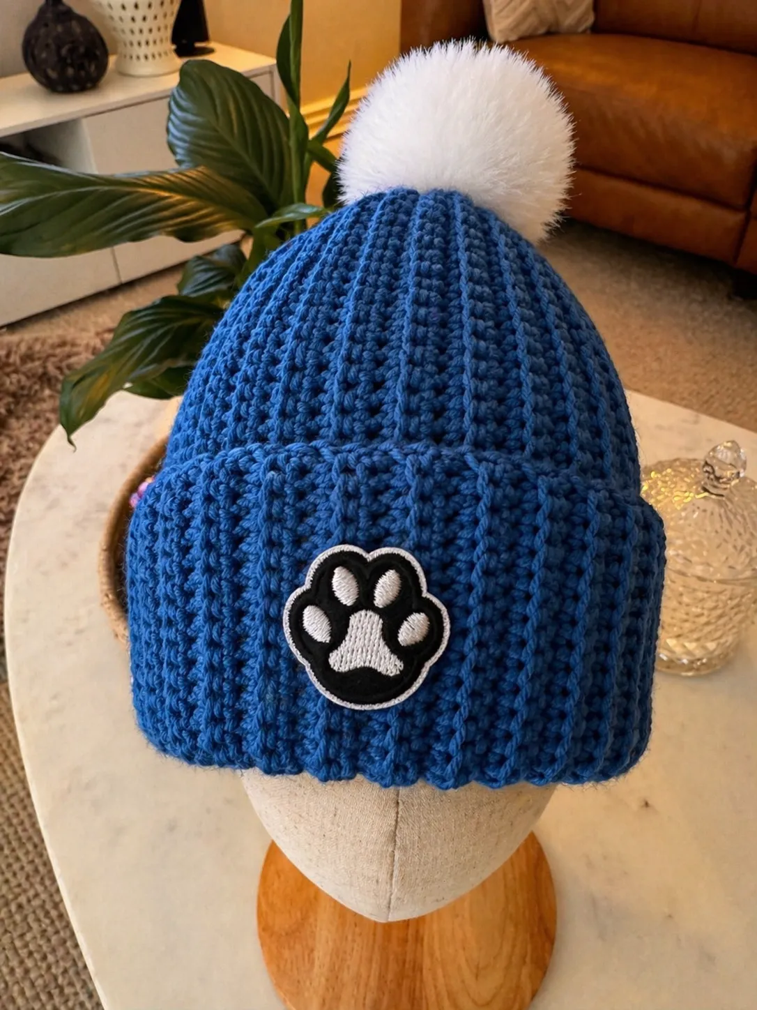 Navy blue crocheted beanie with ribbed texture, featuring a black and white embroidered paw print patch on the front and a fluffy white pom pom on top, displayed on a wooden head form.