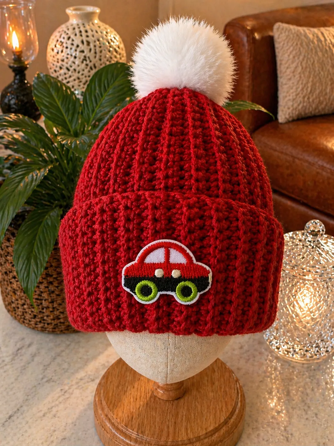 Red crocheted kids beanie with white pom pom and embroidered car patch featuring green wheels, displayed on a wooden head form