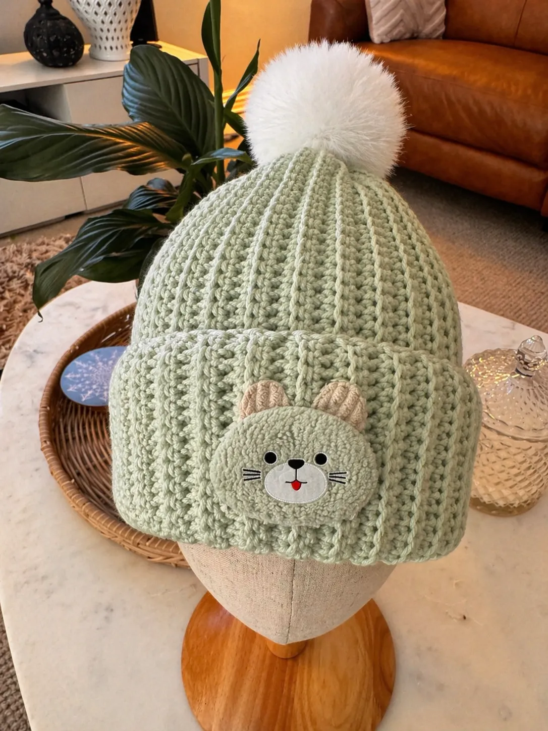 Hand-crocheted sage green beanie with ribbed texture, featuring an embroidered cat face with whiskers and a large white pom pom on top